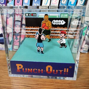 Op de afbeelding: Een heldere acryl display met een retro videogame sc&egrave;ne van Punch-Out!!, met gepixelde karakters die boksen op een blauw platform. De speltitel wordt weergegeven in oranje en blauw. De achtergrond bevat een boekenplank.