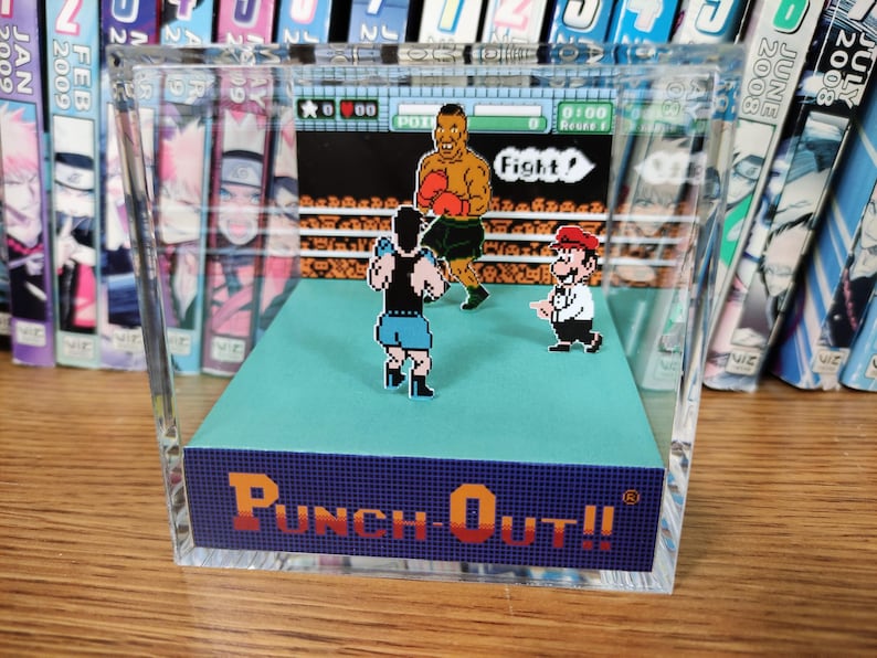 Op de afbeelding: Een heldere acryl display met een retro videogame sc&egrave;ne van Punch-Out!!, met gepixelde karakters die boksen op een blauw platform. De speltitel wordt weergegeven in oranje en blauw. De achtergrond bevat een boekenplank.