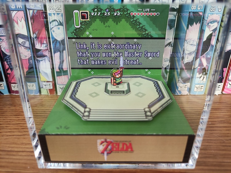 May include: A 3D pop-up diorama of Link from The Legend of Zelda video game series. Link is standing on a platform with a green background. The text "Link, it is extraordinary that you won the Master Sword that makes evil retreat..." is displayed in a speech bubble above Link. The diorama is displayed in a clear acrylic case.