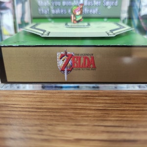 May include: A clear acrylic display case with a green base and a gold-colored bottom panel. The panel features the text "THE LEGEND OF ZELDA A LINK TO THE PAST" in a black font with a red sword graphic.