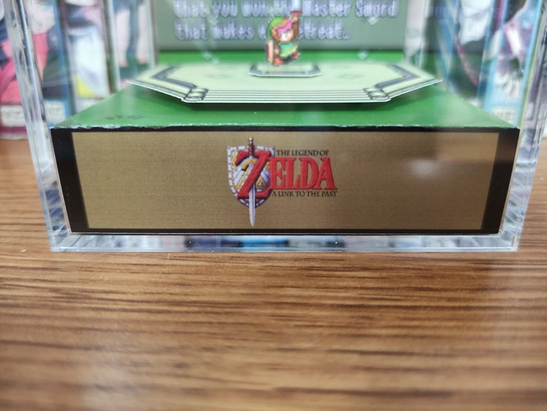 May include: A clear acrylic display case with a green base and a gold-colored bottom panel. The panel features the text "THE LEGEND OF ZELDA A LINK TO THE PAST" in a black font with a red sword graphic.