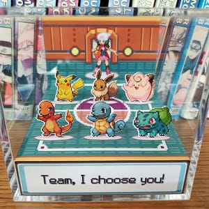 Puede incluir: Bloque acr&iacute;lico transparente con una escena pixelada de Pok&eacute;mon, incluyendo Pikachu y Eevee, y un entrenador. El texto "Team, I choose you!" est&aacute; en la base. Mangas rodean el bloque. La escena evoca un videojuego.