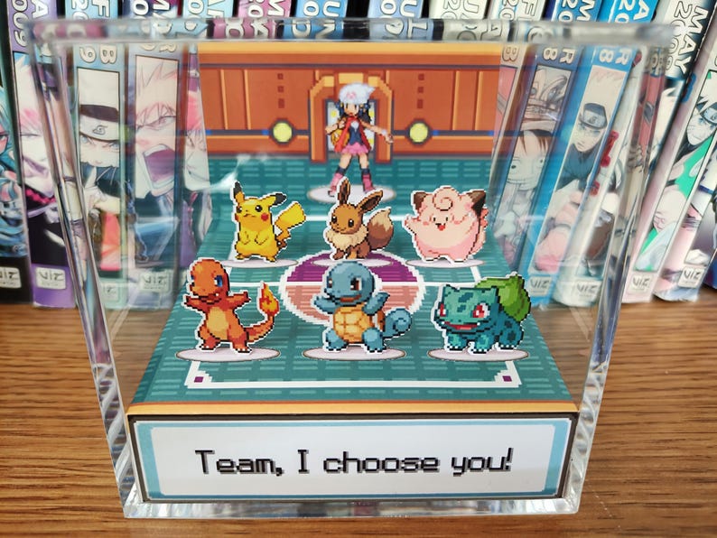 Puede incluir: Bloque acr&iacute;lico transparente con una escena pixelada de Pok&eacute;mon, incluyendo Pikachu y Eevee, y un entrenador. El texto "Team, I choose you!" est&aacute; en la base. Mangas rodean el bloque. La escena evoca un videojuego.