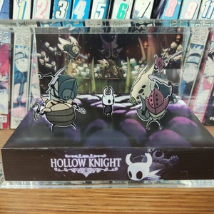 May include: A clear acrylic display case with a black background featuring characters from the video game Hollow Knight. The case has a black base with the words "Hollow Knight" printed in white. The characters include the Knight, a small, white, bug-like creature with a glowing mask, and several other characters from the game.
