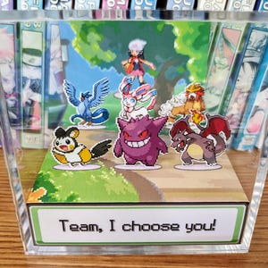 Puede incluir: Una pantalla acr&iacute;lica transparente con un diorama de varios personajes de Pok&eacute;mon. La escena incluye un entrenador y varios Pok&eacute;mon, con el texto "Team, I choose you!" en la base. El fondo representa un paisaje herboso.