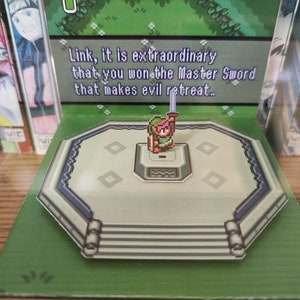 May include: A pop-up book featuring a pixelated character holding a sword. The text reads "Link, it is extraordinary that you won the Master Sword that makes evil retreat..."