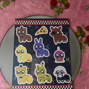 Puede incluir: Una hoja de pegatinas con personajes de dibujos animados de varios colores. Las pegatinas incluyen un oso marrón, un conejo morado, un zorro rojo, un pollito amarillo, un conejo verde, una marioneta blanca, una porción de pizza y una magdalena. Las pegatinas están sobre un fondo azul oscuro.