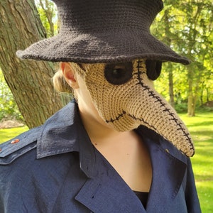May include: A person wearing a brown crocheted plague doctor mask with a black crocheted hat. The mask has a long, pointed nose and eye holes with black fabric around them.