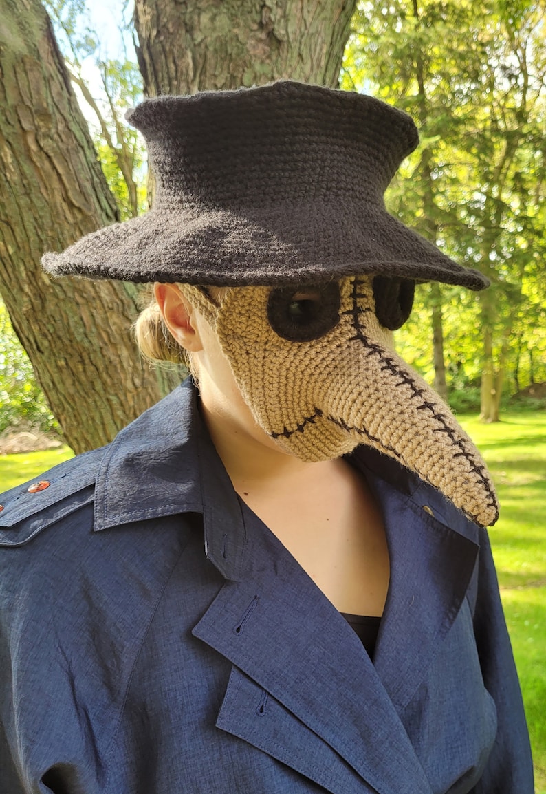May include: A person wearing a brown crocheted plague doctor mask with a black crocheted hat. The mask has a long, pointed nose and eye holes with black fabric around them.