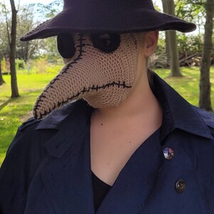 May include: A person wearing a black hat and a brown crocheted plague doctor mask. The mask has a long, pointed beak and two eye holes. The person is wearing a dark blue coat.