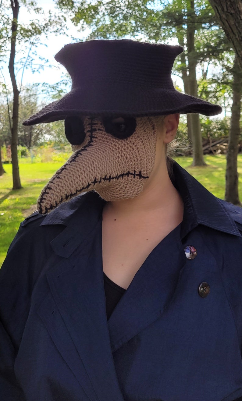 May include: A person wearing a black hat and a brown crocheted plague doctor mask. The mask has a long, pointed beak and two eye holes. The person is wearing a dark blue coat.