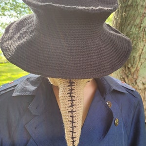 May include: A black crocheted hat with a wide brim and a beige crocheted plague doctor mask.
