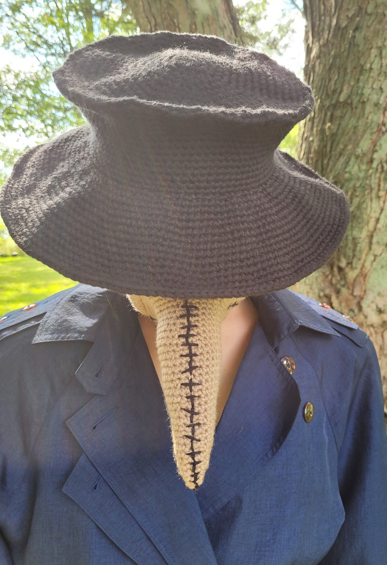 May include: A black crocheted hat with a wide brim and a beige crocheted plague doctor mask.