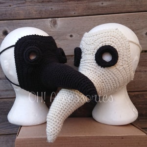 May include: Two crocheted plague doctor masks, one black and one white, with long beaks. The masks are displayed on styrofoam heads.