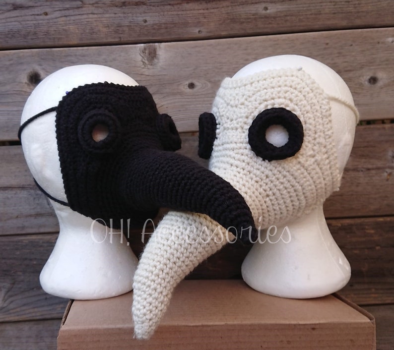 May include: Two crocheted plague doctor masks, one black and one white, with long beaks. The masks are displayed on styrofoam heads.