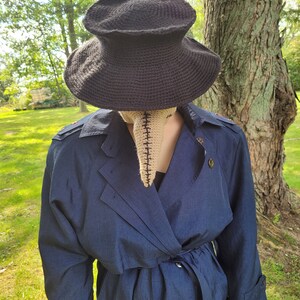 May include: A person wearing a dark blue trench coat with a black crocheted hat and a beige crocheted mask with a long nose.