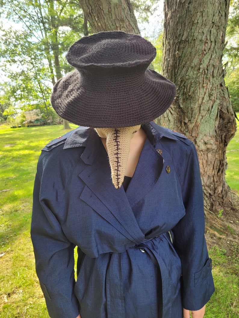 May include: A person wearing a dark blue trench coat with a black crocheted hat and a beige crocheted mask with a long nose.