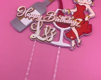May include: A cake topper featuring a cartoon character pouring wine into a glass. The topper includes the words "Happy Birthday Liz" in gold and pink lettering.