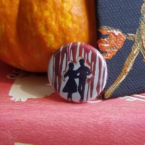 May include: A round button with a red and white striped background featuring a silhouette of a couple dancing. The button is sitting on top of two red and green books with the text "L.M. Montgomery" and "Anne" visible.