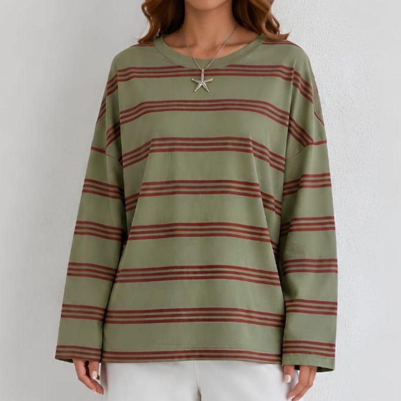May include: An olive green long-sleeve shirt featuring horizontal dark red stripes and a crew neckline. A silver-toned starfish pendant hangs from a delicate chain, resting against the center of the chest over the striped fabric pattern.
