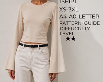 Puede incluir: Una camiseta beige de manga larga con mangas acampanadas y cuello redondo. El texto a la derecha dice: BELL SLEEVE TSHIRT, XS-3XL, A4-A0-LETTER, PATTERN+GUIDE, DIFFUCULTY LEVEL y dos estrellas. La camiseta se combina con vaqueros blancos y un cinturón negro.