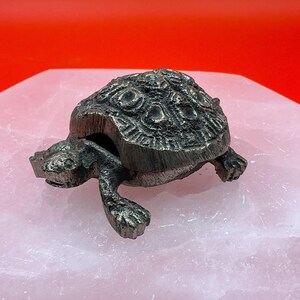 May include: A detailed, antique-style metal turtle figurine with a textured shell and outstretched legs. The turtle is a dark, metallic color and sits on a light pink surface against a red background.