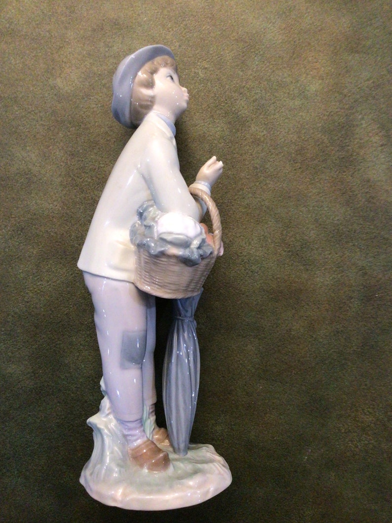 May include: A porcelain figurine of a boy wearing a blue hat and a blue and white jacket, holding a basket of fruit and an umbrella.