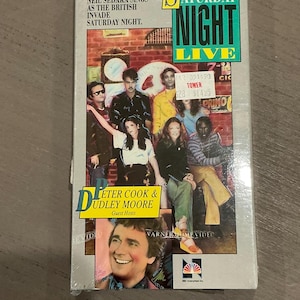 May include: A VHS tape cover for an episode of Saturday Night Live featuring Neil Sedaka, Peter Cook, and Dudley Moore. The cover is white with blue and red text. The image shows the cast of Saturday Night Live.