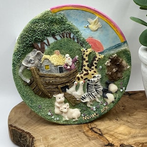 Könnte beinhalten: Ein Keramikteller mit einer 3D-Darstellung der Arche Noah mit Tieren, darunter eine Giraffe, ein Zebra, ein Elefant und Schweine. Die Arche ist von einem grünen Feld mit Blumen und einem Regenbogen am Himmel umgeben. Der Teller ist mit den Worten "Noah's Ark" beschriftet.