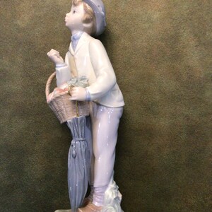 May include: A porcelain figurine of a boy holding a basket of fruit and an umbrella. He is wearing a white jacket, blue pants, and a blue hat. The figurine is standing on a base with green grass and flowers.