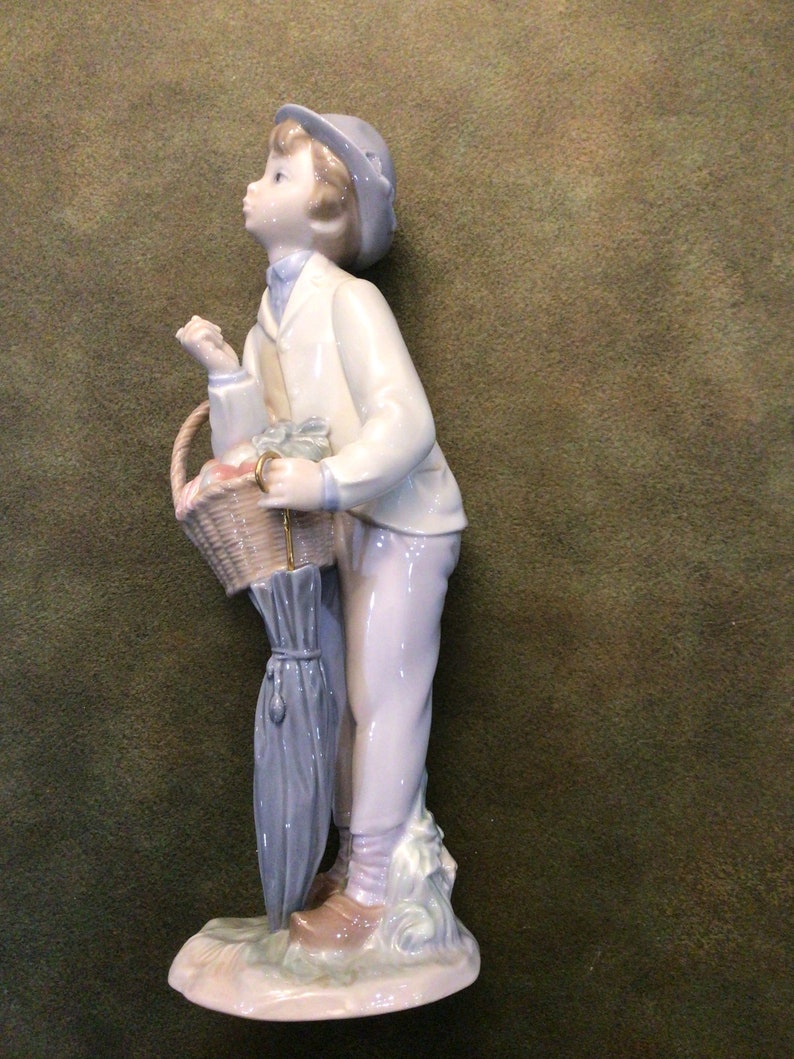 May include: A porcelain figurine of a boy holding a basket of fruit and an umbrella. He is wearing a white jacket, blue pants, and a blue hat. The figurine is standing on a base with green grass and flowers.