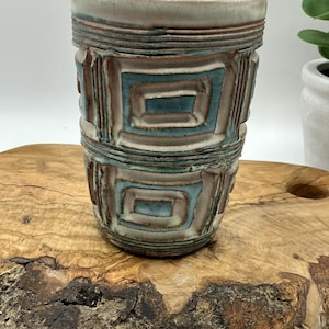 May include: A cylindrical ceramic tumbler featuring a textured surface with raised geometric rectangular patterns. The vessel displays a color palette of muted white, earthy brown, and soft teal blue glazes, with horizontal ridges encircling the top.