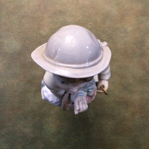 May include: A porcelain figurine of a child wearing a grey helmet and holding a rope. The figurine is painted in shades of blue, pink, and white.