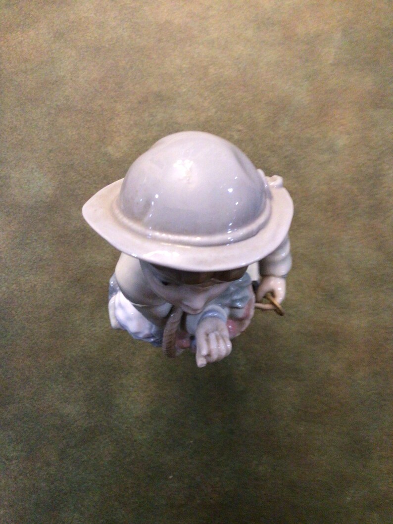 May include: A porcelain figurine of a child wearing a grey helmet and holding a rope. The figurine is painted in shades of blue, pink, and white.