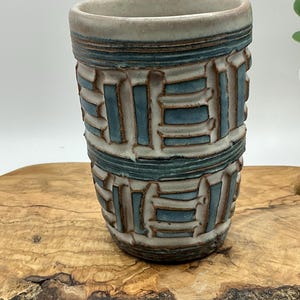 May include: A cylindrical ceramic tumbler featuring a textured surface with a repeating geometric pattern. The design consists of raised white clay ridges interspersed with recessed blue-toned glaze, creating a tactile, layered appearance.