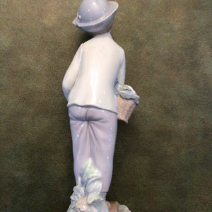 May include: A porcelain figurine of a boy wearing a blue hat and carrying a basket of vegetables. The figurine is standing on a base with green and white details.