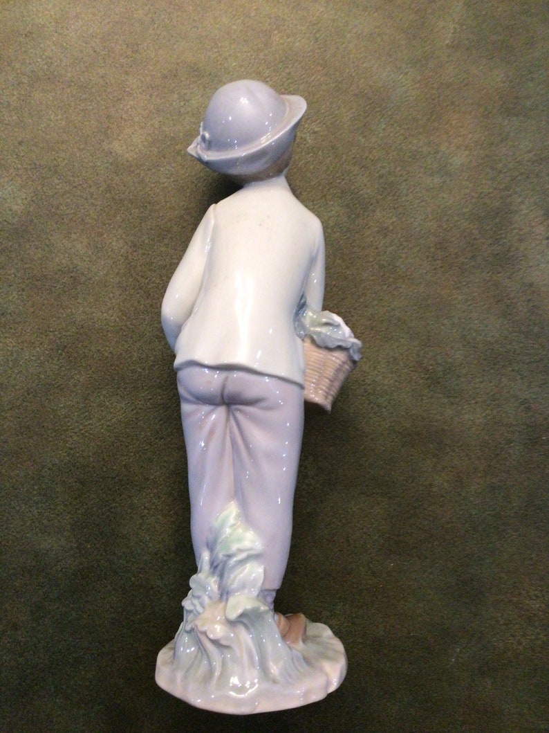 May include: A porcelain figurine of a boy wearing a blue hat and carrying a basket of vegetables. The figurine is standing on a base with green and white details.