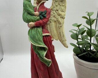 Może przedstawiać: Ceramiczna figurka anioła trzymającego dziecko. Anioł ma na sobie czerwoną sukienkę z zielonym szalem i duże, białe skrzydła. Dziecko ma na sobie zieloną sukienkę i blond włosy.
