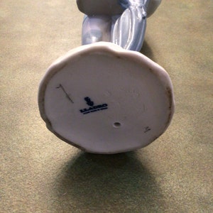 May include: A white porcelain figurine of a girl carrying a wicker basket and an umbrella. The figurine has a blue and white floral pattern and is marked with the Lladro logo on the bottom.