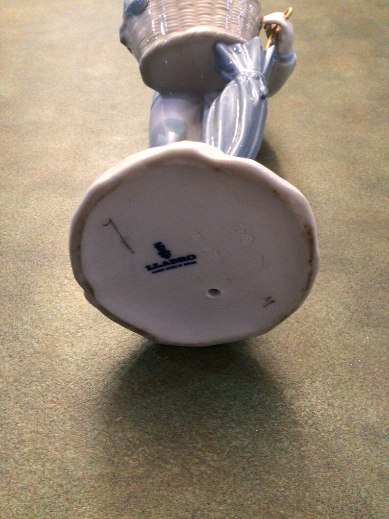 May include: A white porcelain figurine of a girl carrying a wicker basket and an umbrella. The figurine has a blue and white floral pattern and is marked with the Lladro logo on the bottom.
