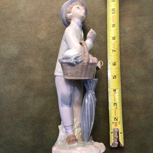 May include: A porcelain figurine of a boy wearing a blue hat and holding a basket and umbrella. The figurine is standing on a base with a light blue and white design.