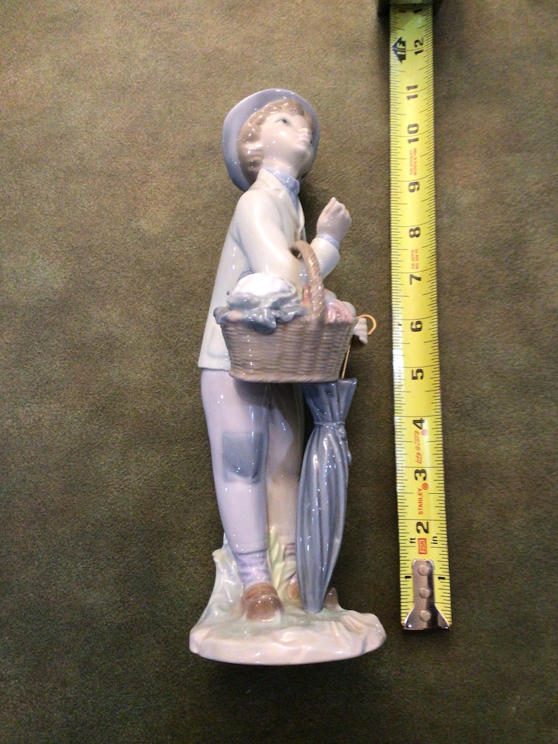 May include: A porcelain figurine of a boy wearing a blue hat and holding a basket and umbrella. The figurine is standing on a base with a light blue and white design.
