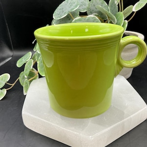 May include: A bright green ceramic mug with a rounded handle. The mug is sitting on a white, hexagonal coaster.