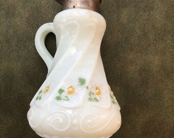 May include: A white, antique glass creamer with a silver metal lid. The creamer has a handle and is decorated with painted flowers and green leaves. The glass has a swirled design. The creamer is on a green background.
