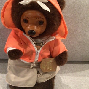 Op de afbeelding: Een bruine teddybeer die een feloranje capuchontrui draagt met een witte strik. De beer draagt een beige broek en heeft een houten gezicht. Het label op de beer luidt "Mijn prinsessen vriendinnen".