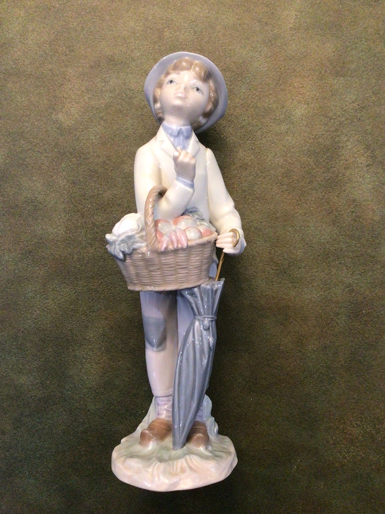 May include: A porcelain figurine of a young person wearing a blue hat and holding a basket of fruit and an umbrella.