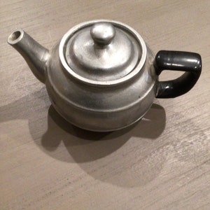 May include: A silver teapot with a black handle and a lid. The teapot is sitting on a light brown wooden surface.