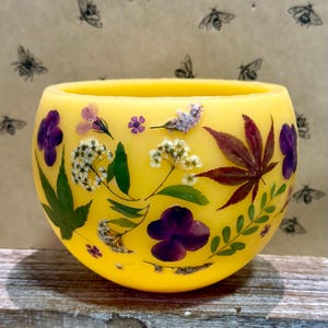 May include: A round, yellow candle holder adorned with pressed flowers and leaves. The candle holder features purple pansies, white blossoms, and a deep red maple leaf. The background has a pattern of black bee illustrations.
