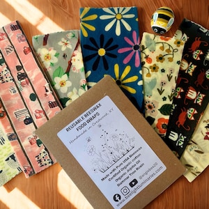 May include: A collection of reusable beeswax food wraps with various colorful patterns, including floral, sheep, and geometric designs. A small box with a label that reads "REUSABLE BEESWAX FOOD WRAPS" is also present, along with a decorative bee figurine.