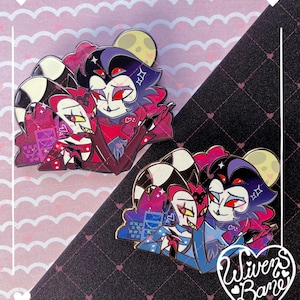 May include: Two enamel pins featuring cartoon characters in shades of red, purple, and blue. The pins depict two characters embracing, with a moon and stars in the background. The pins have gold-colored metal borders and are set against a pink and black patterned background.